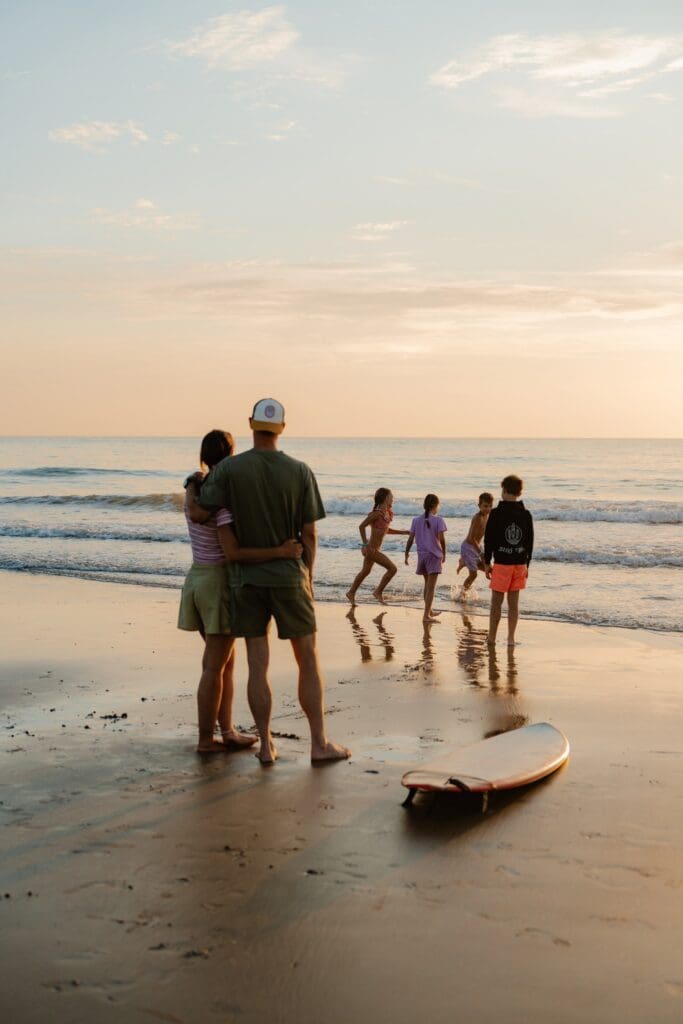 family watching kids play in ocean at sunset with surfboard